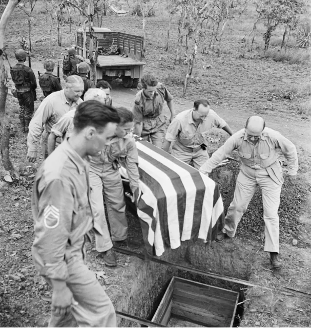 Looking down on an open grave in red earth among thin trees: men in khaki shirts lower a coffin draped in the American flag; an American sergeant stands at the left.