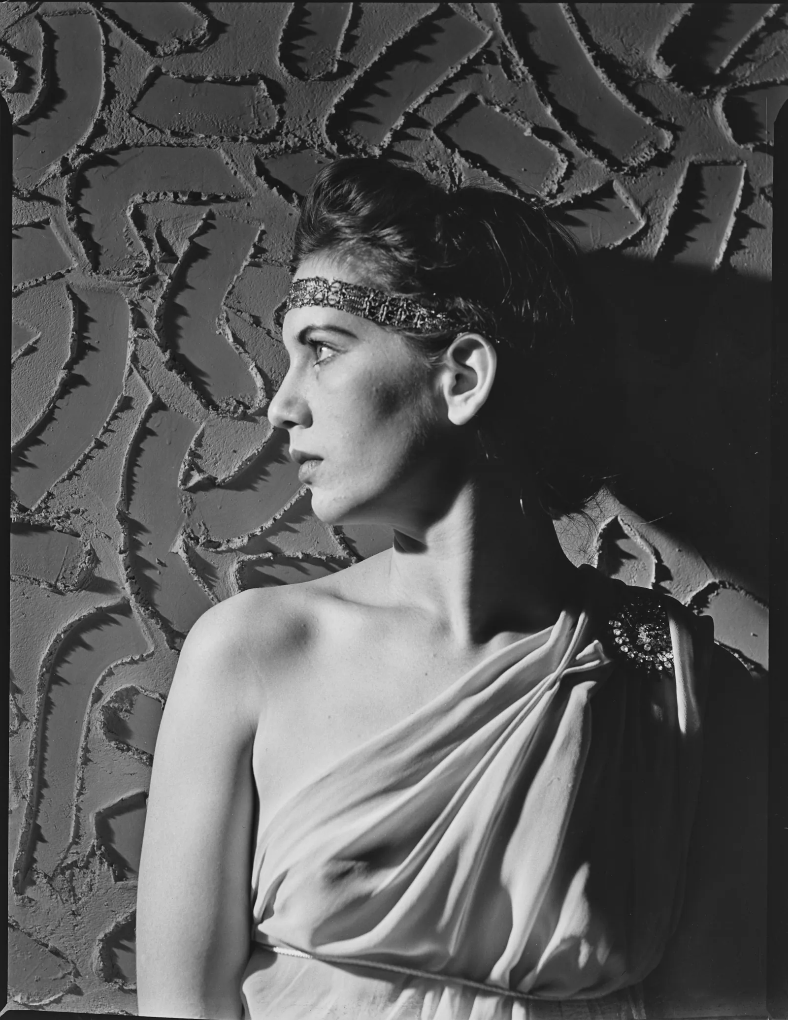 A studio portrait of a young woman in left profile, her dark hair swept up under a jewelled headband, one shoulder bare and the other draped in a pale gown pinned with a brooch, her shadow on a patterned wall.