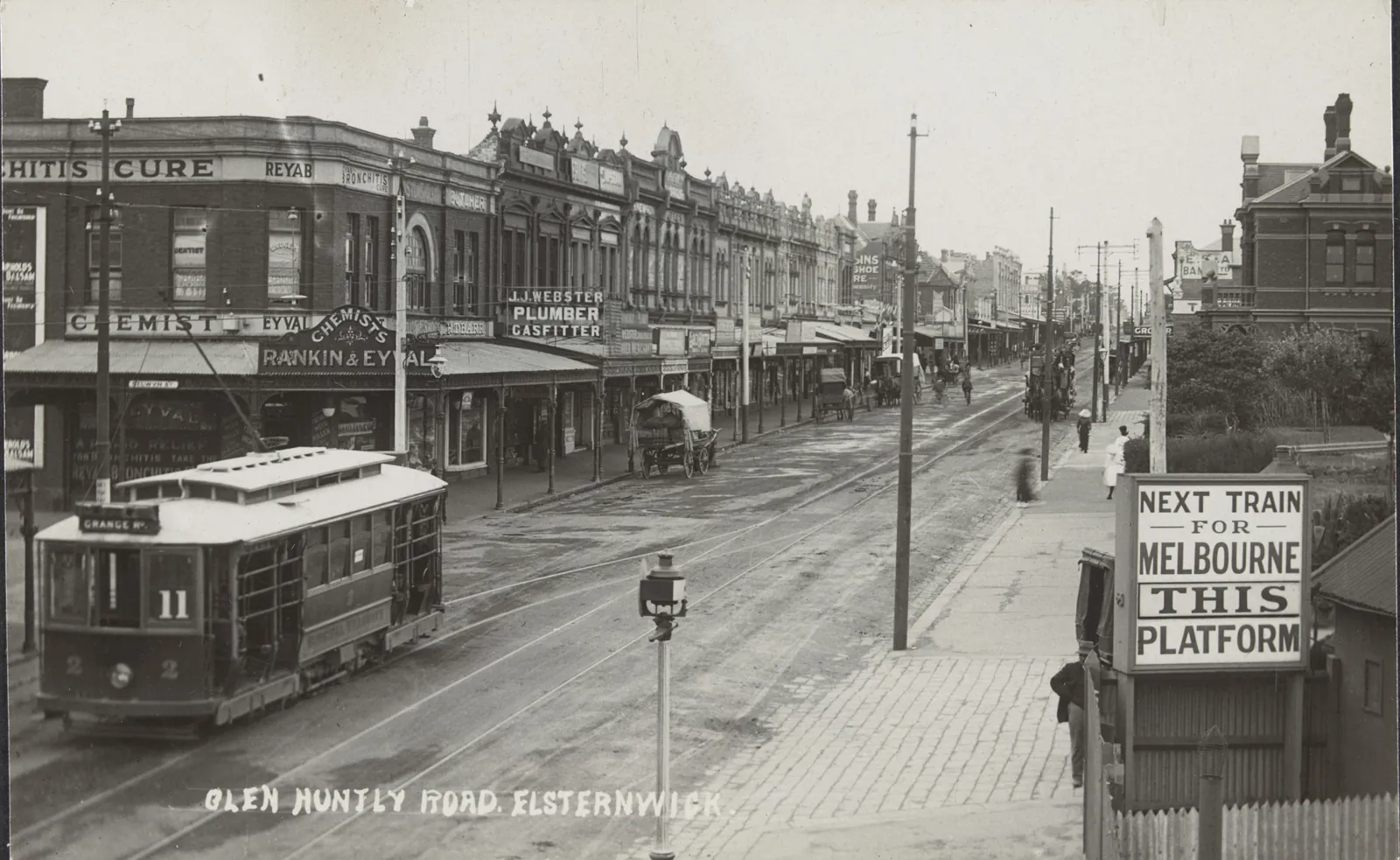 A postcard view along Glen Huntly Road, Elsternwick: an electric tram, number 11, on the left, two-storey shops with verandahs receding along the road, a horse and cart, telegraph poles, and on the right a sign on the station fence, “Next train for Melbourne this platform”.