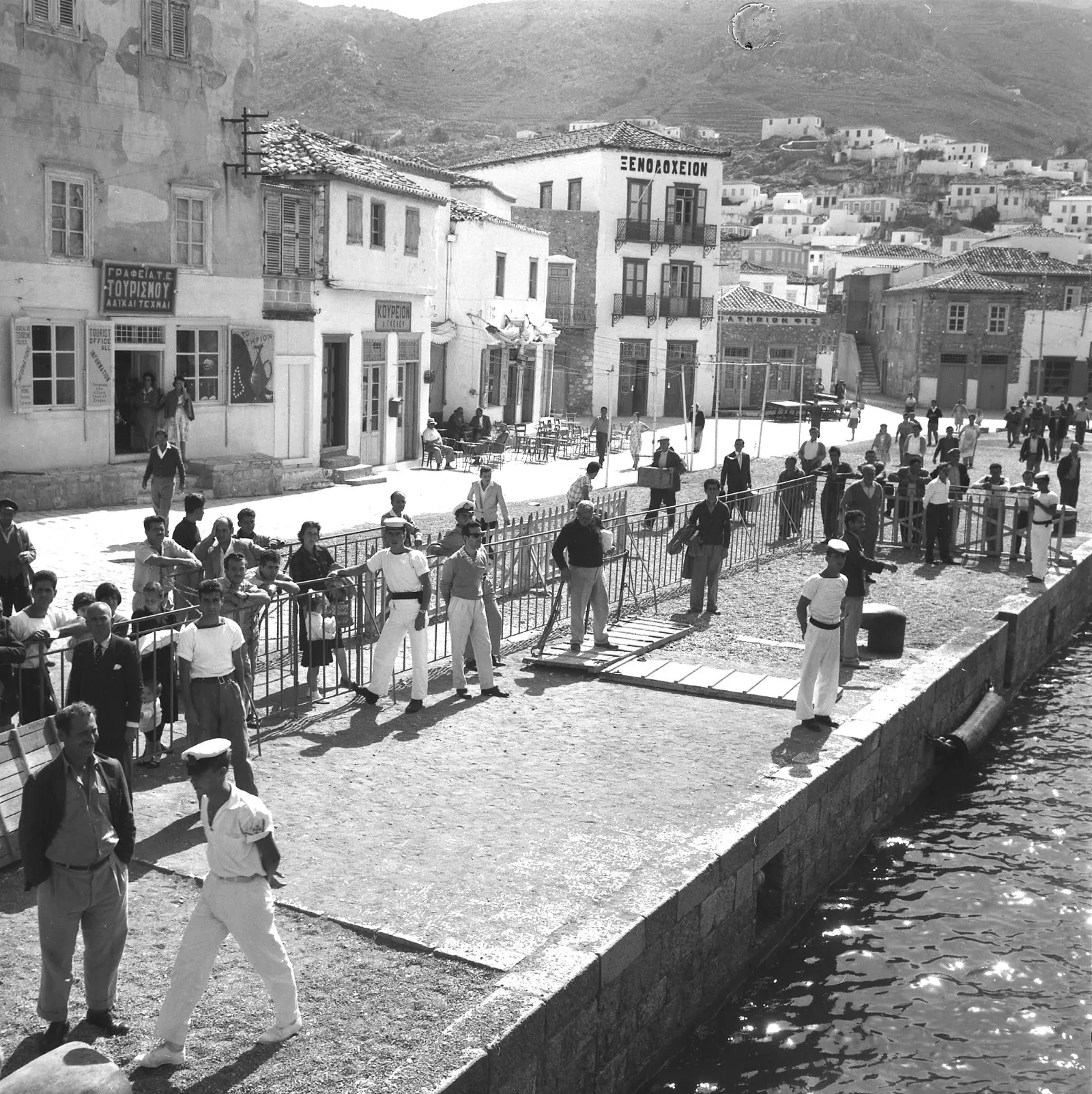 Seen from an arriving boat, a stone quay with a crowd waiting behind an iron barrier, sailors in white among them; behind, a tourist office, a barber’s, café tables, a hotel with iron balconies, and white houses climbing a bare hill.